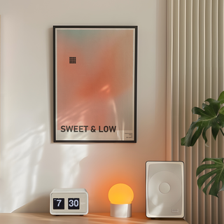 Urban typography poster with gradient peach design and text 'SWEET & LOW'. Displayed in a black wooden frame. Placed in a minimal room next to a plant and lamp.
