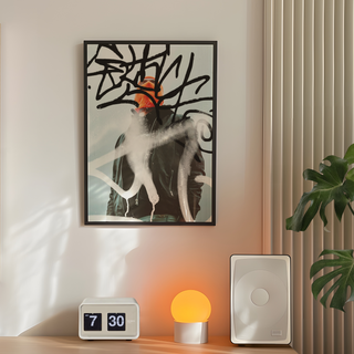 Urban photography of man wearing an orange beanie and black jacket with abstract art graffiti elements on a light blue background. Displayed in a black wooden frame placed in a bedroom with lamp and plant.