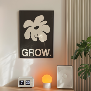 Framed artwork featuring a white abstract leaf design and the word 'GROW.' on a black background, displayed in a premium black wooden frame. Placed on bedroom wall next to plant, speakers and lamp.