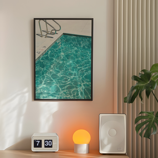 Framed photograph of a turquoise blue pool with clear water, displayed in a premium black wooden frame. Displayed on a bedroom wall next to plant, lamp and speakers.