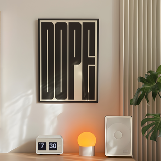 Bold black and white typography poster with the word 'DOPE' in bold, black letters on a white background. Displayed in a black premium wood frame. Placed on the wall in a bedroom next to speakers, lamp and plant.