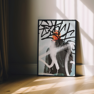 Urban photography of man wearing an orange beanie and black jacket with abstract art graffiti elements on a light blue background. Displayed in a black wooden frame.