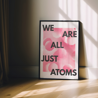 Framed poster with 'We are all just atoms' text on a pink background, placed on a wooden floor with sunlight casting shadows.