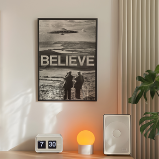 Black and white photography surreal poster with a UFO over a landscape and the word 'BELIEVE' below. Displayed in a black wooden frame next to lamp, plant and speakers in bedroom.