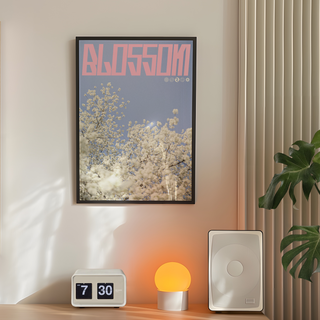 Lilac and pink graphic design poster with cherry blossoms and 'Blossom' text. Displayed in a premium wooden black frame. Placed on the wall in a bedroom next to plant, speakers and lamp.