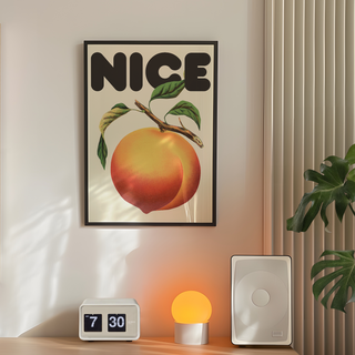 Framed artwork featuring a vintage-style peach and leaves with the word 'NICE' in bold text on a beige background, displayed in a premium black wooden frame. Placed in bedroom next to plant, speakers and lamp.