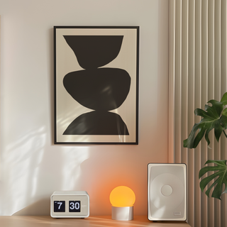 Minimalist black and white abstract poster with three bold black shapes stacked against a clean white background, showcased in a premium black wooden frame. Placed in bedroom next to plant, lamp and speakers. 
