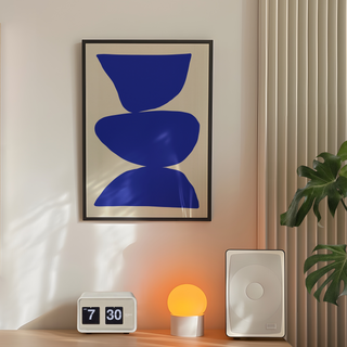 Cobalt blue abstract art with blue shapes on a beige background. Displayed in a black wooden frame. Placed in a minimalistic bedroom next to plant, speakers and lamp. 