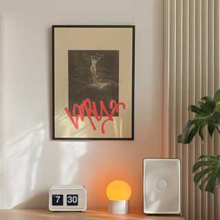 Vintage sepia photograph of nude lady with red graffiti mark overlaid placed on beige background poster. Displayed in a premium black wooden frame. Placed on a wall in a minimal bedroom next to plant, speakers and lamp. 