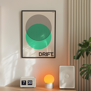 Bold Green circle abstract illustration with black typography poster with the word "drift" written at the bottom. Displayed in a premium wooden black frame.  Placed on a wall in a bedroom next to plant, speakers and lamp. 
