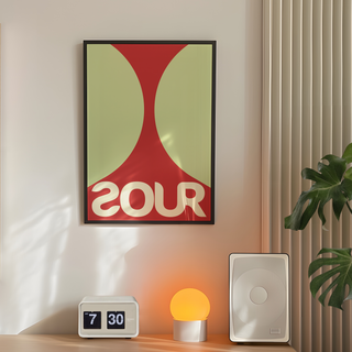 Bold red and green typography poster featuring the word 'SOUR', displayed in a premium black wooden frame. Displayed on a bedroom wall next to speakers, plant and lamp. 
