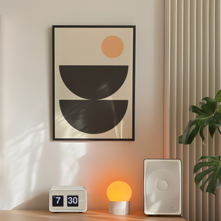 Boho scandi style abstract art print with black semi circle shapes and a peach circle on a beige background. Displayed in a premium wood black frame.  Placed in a bedroom next to plant, speakers and lamp. 