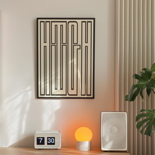 Bold black and white typography poster featuring the word 'HIGH' filling the entire page, presented in a premium black wooden frame. Displayed on the wall in a bedroom next to plant, speaker and lamp. 